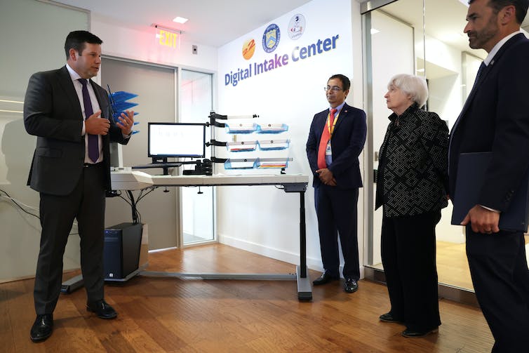 Three men in suits and Janet Yellen stand around a computer and a sign on the wall reading 'digital intake center.'