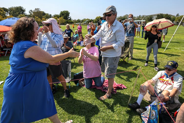 Two people reach their hands out to each other to shake hands in a grassy area with other people around.