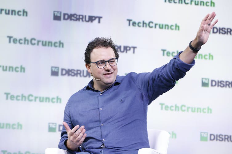 Man in blue button-down shirt gesticulates with one arm outstretched against a backdrop with the words TechCrunch and DISRUPT