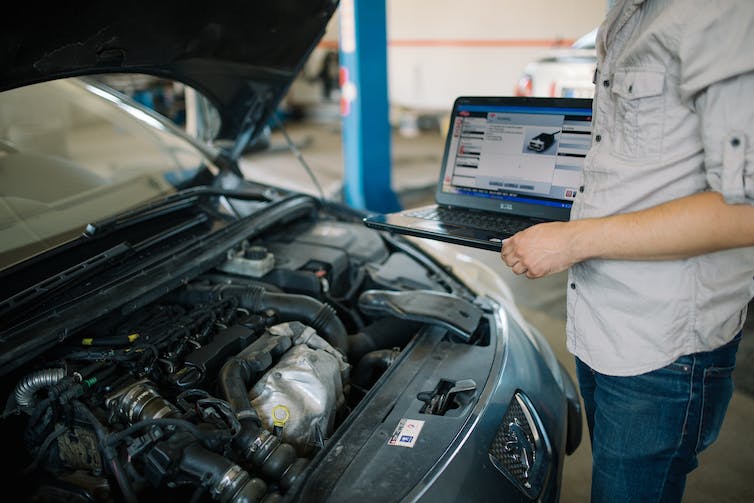 torso of a person holding a laptop computer in front of a car with its hood raised showing the engine compartment