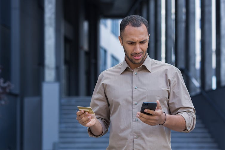 Frustrated and upset man outside office building looking at his smartphone and holding a bank card