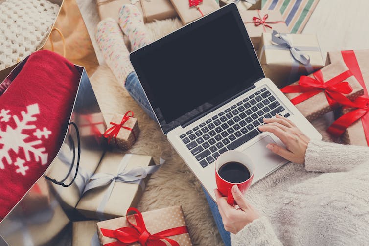 A person sits at a laptop with a coffee surrounded by festive packages.