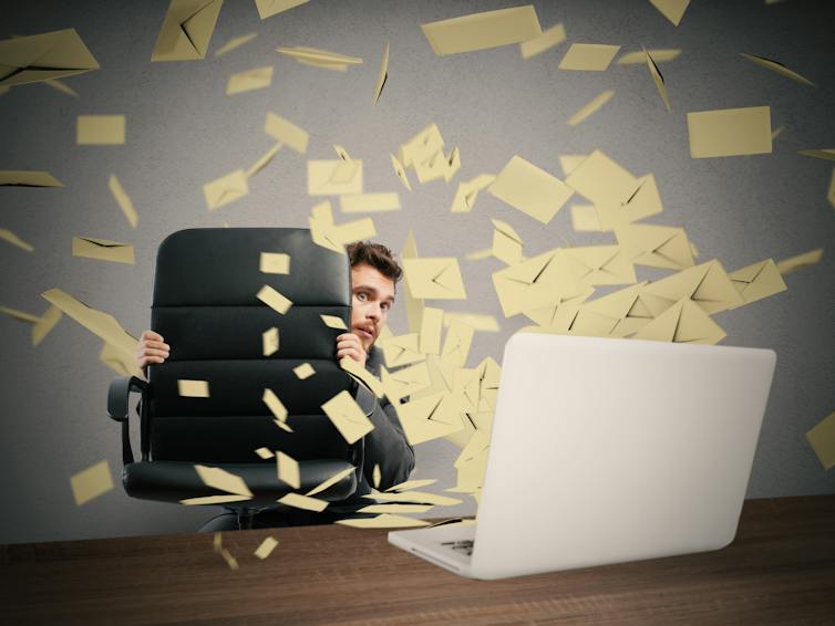 Emails seems to explode out of laptop as man hides behind office chair