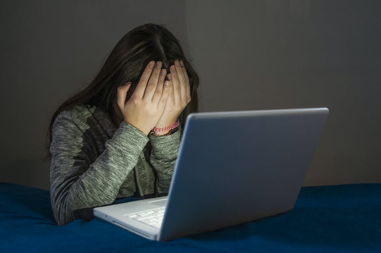 young woman with face in her hands sits in front of a laptop