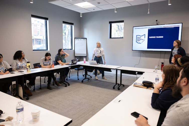 Young people sit around a table, and two young people, both wearing white T-shirts, stand near a screen that says 'Canvass training'