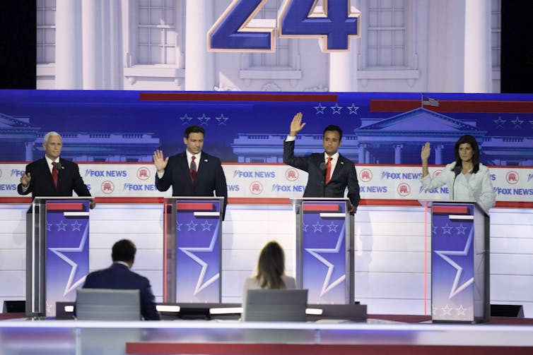 Four people stand behind lecterns on a stage.