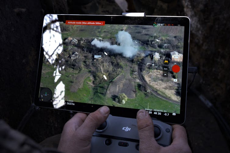 a pair of hands hold a small video monitor displaying an aerial view of a landscape