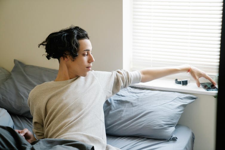 A teenage boy lies on a bed, reaching for his phone on a shelf behind his head.