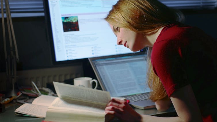 A woman peers into a book while seated at her desk, which has both a desktop and a laptop computer.