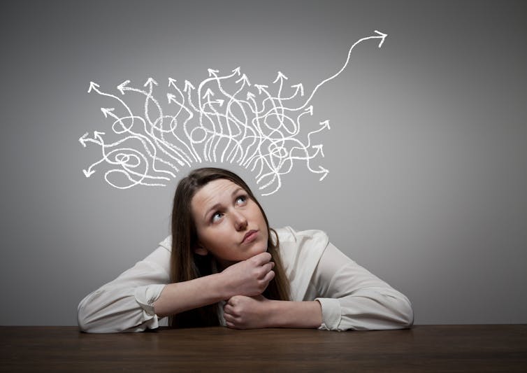 Girl thinking with arms resting on a table, arrows in different directions above her head