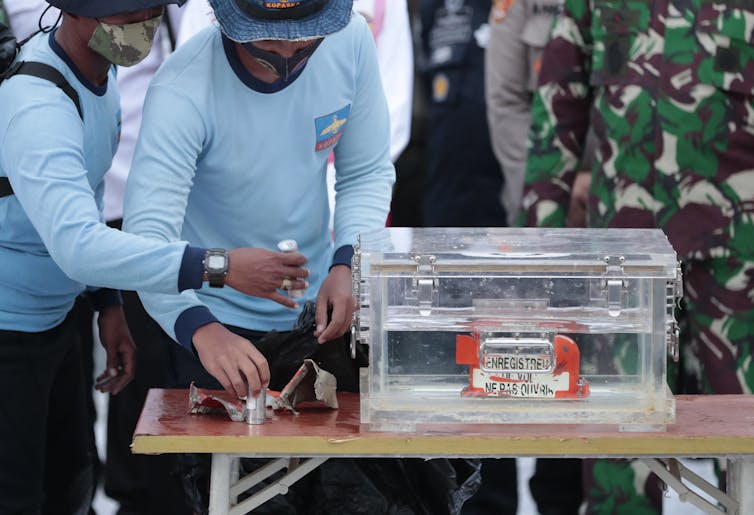 two men in light blue uniforms stand next to a clear box on a table