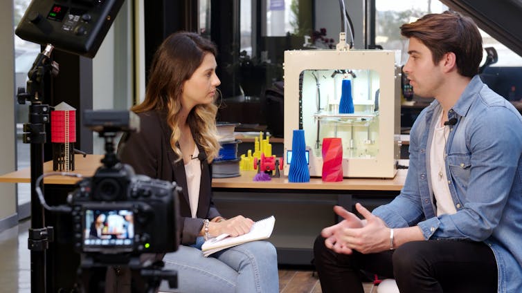 Video camera points at woman interviewing a man in a tech setting