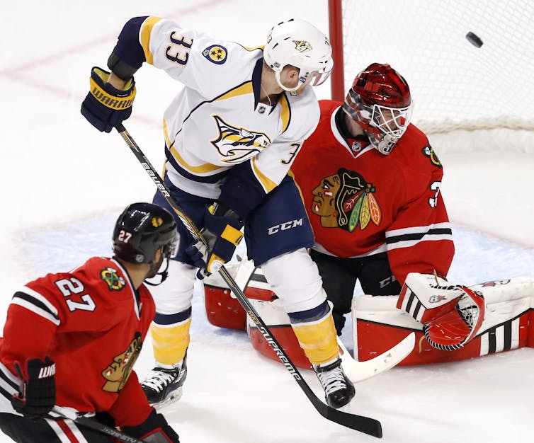 Hockey players watch a puck fly over the shoulder of a goalie after being deflected.