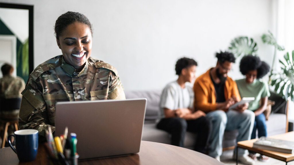 A smiling military service member uses a laptop at a table while three people in the background sit on a couch and view a tablet together.