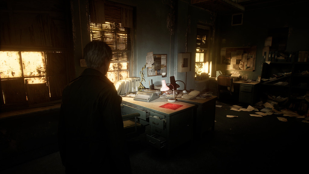 This images shows James standing in an office-like environment. Papers are strewn over the floor, with the setting illuminated by a glimmer of light from three sets of grimy windows.