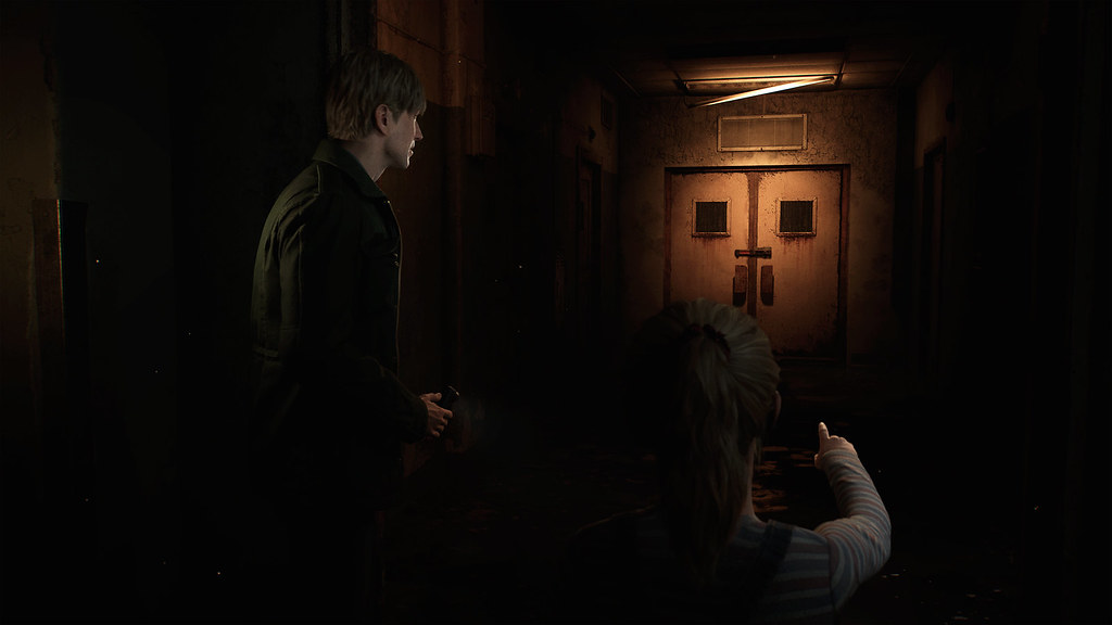 In this image, James stands to the left of the screen, staring at a rusted set of double doors at the end of a dark corridor. His gaze is directed that way by the pointed finger of a young girl who stands alongside him.