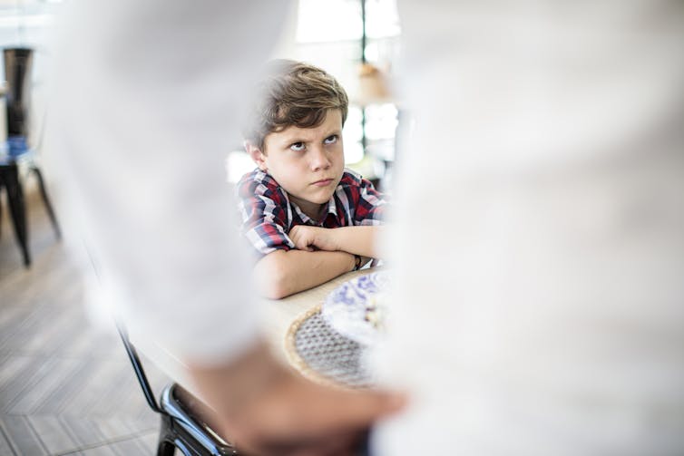 View between the arm of a person with their hands on their hips, focusing on a child sitting at table with a glare