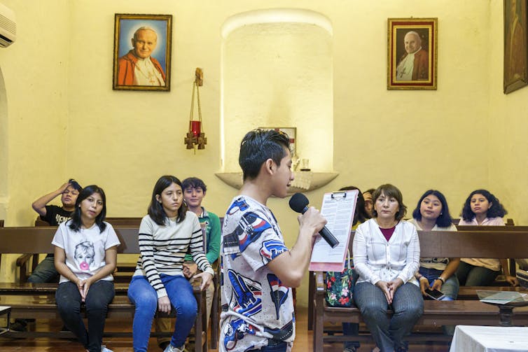 A teenage boy speaks on the microphone as several other young people seated in pews listen to him.