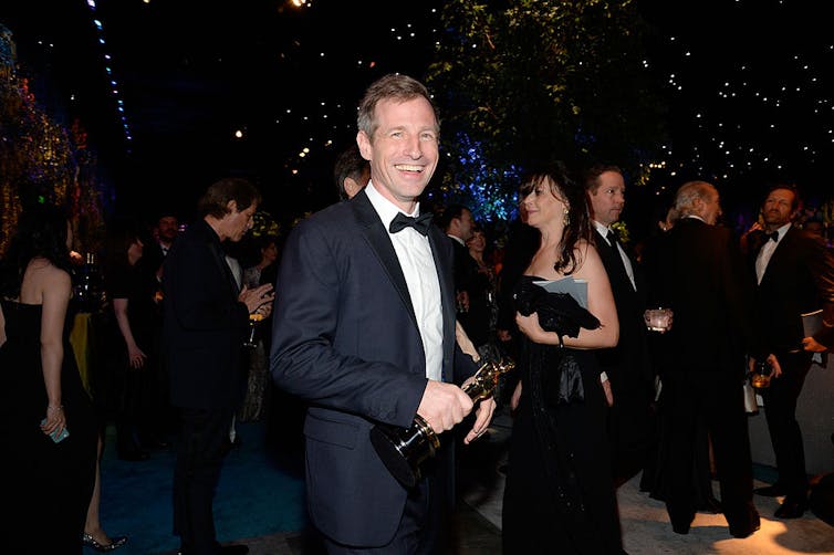 Young man wearing tuxedo smiles as he holds a gold statuette.