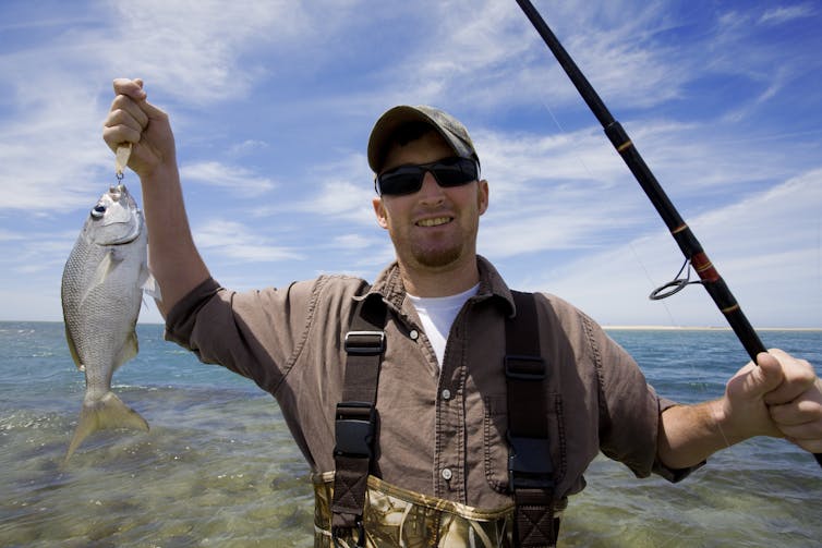 a man wearing sunglasses and a hat holds up a fish and a fishing rod