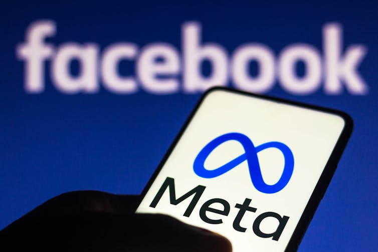 A hand holds a cellphone with the Meta logo on it in front of a screen displaying the word facebook