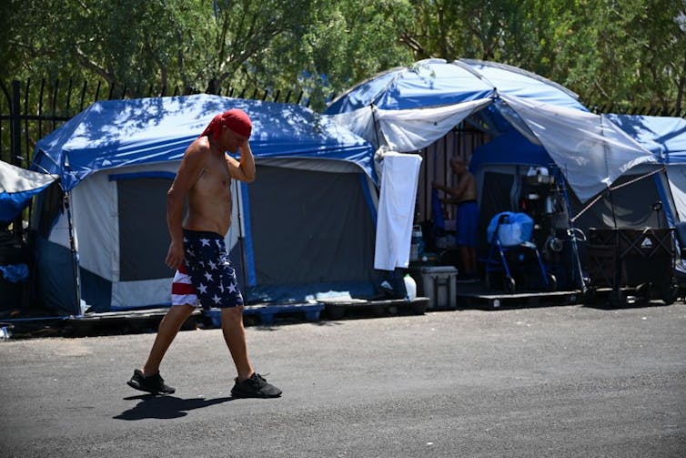 Shirtless man wearing red bandana and stars and stripes shorts walks by a row of tents.