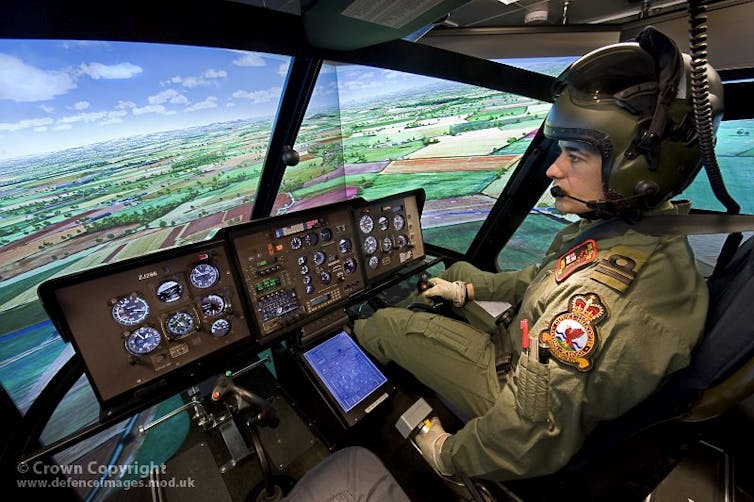 a man wearing a uniform and helmet sits at aircraft controls and looks at displays screens showing simulated landscape