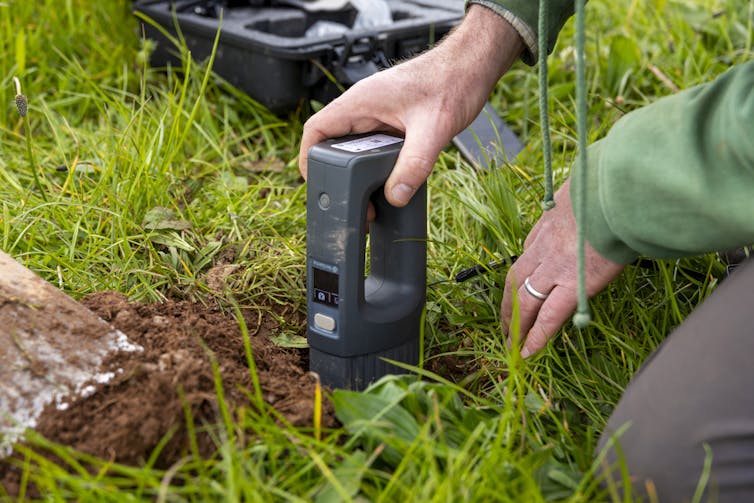 close up shot of person's hands holding small black computer kit by soil and grass