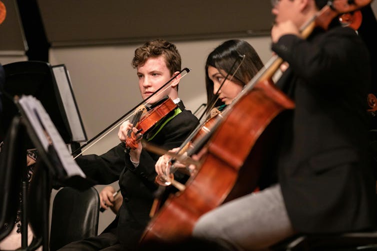 Three students play in an orchestra. Two play a violin, one is on the cello.