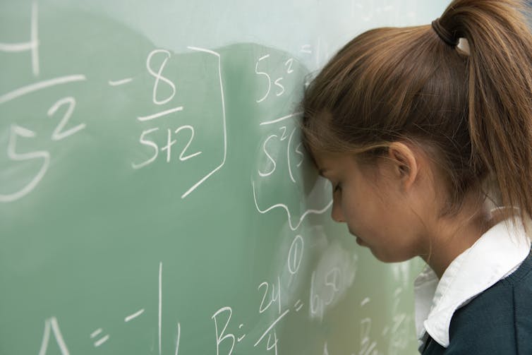 A girl rests her head on a chalkboard, seemingly exasperated over her inability to do a math problem.