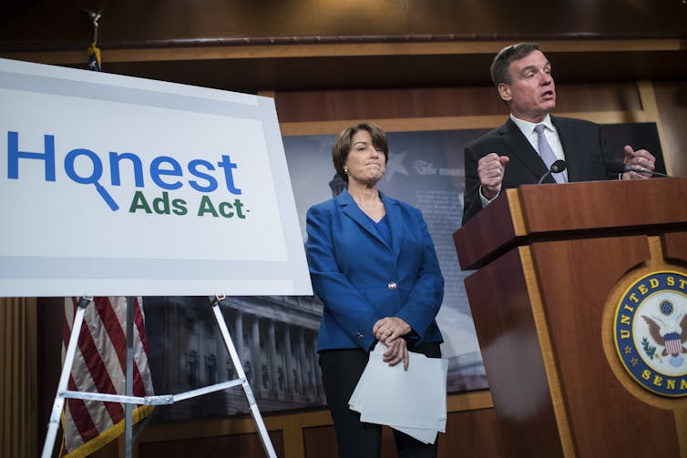 A man in a business suit stands at a podium while a woman in a business suit stands between the podium and a sign on an easel