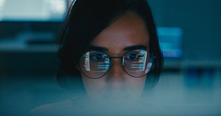 The glare of a laptop screen reflects in a woman's eyeglasses.