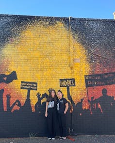 Two First Nations researchers standing in front of red, black and yellow mural featuring silhouettes of protestors holding signs that read land back and sorry means you don't do it again.