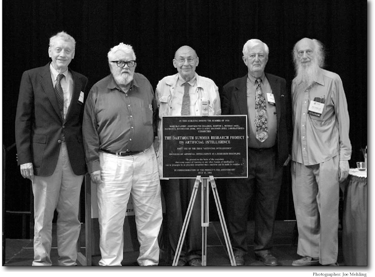 Five elderly men standing on a stage in front of a commemorative plaque