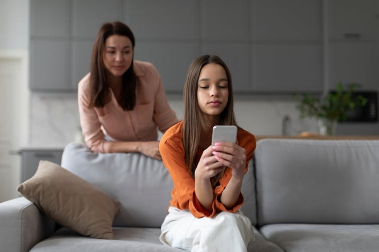 Mother looking over shoulder of teen daughter sitting on sofa using smartphone