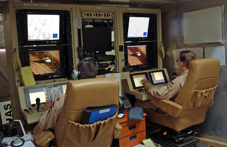 Two men sit at a bank of screens with joysticks