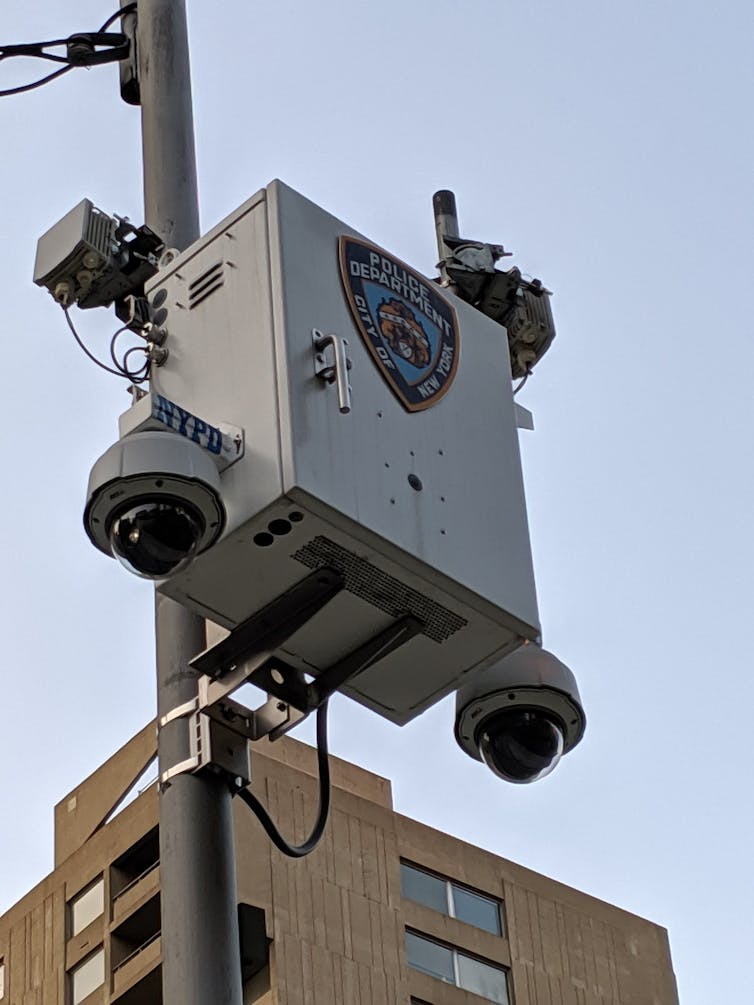 A box mounted on a pole outdoors with several electronic devices attached and two glass semispheres attached at the bottom