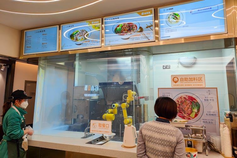 a robotic arm behind a glass wall as two people stand in front of the glass watching