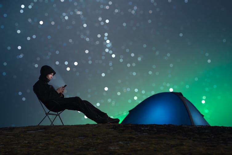 A man leans on a camping chair outdoors, looking at his phone, with an aurora in the background.