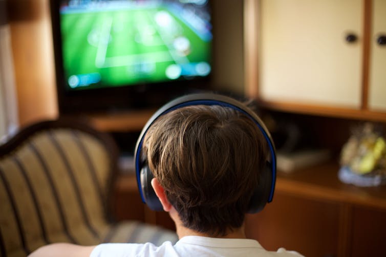 Back of a boy's head wearing headphones and looking at a TV screen.