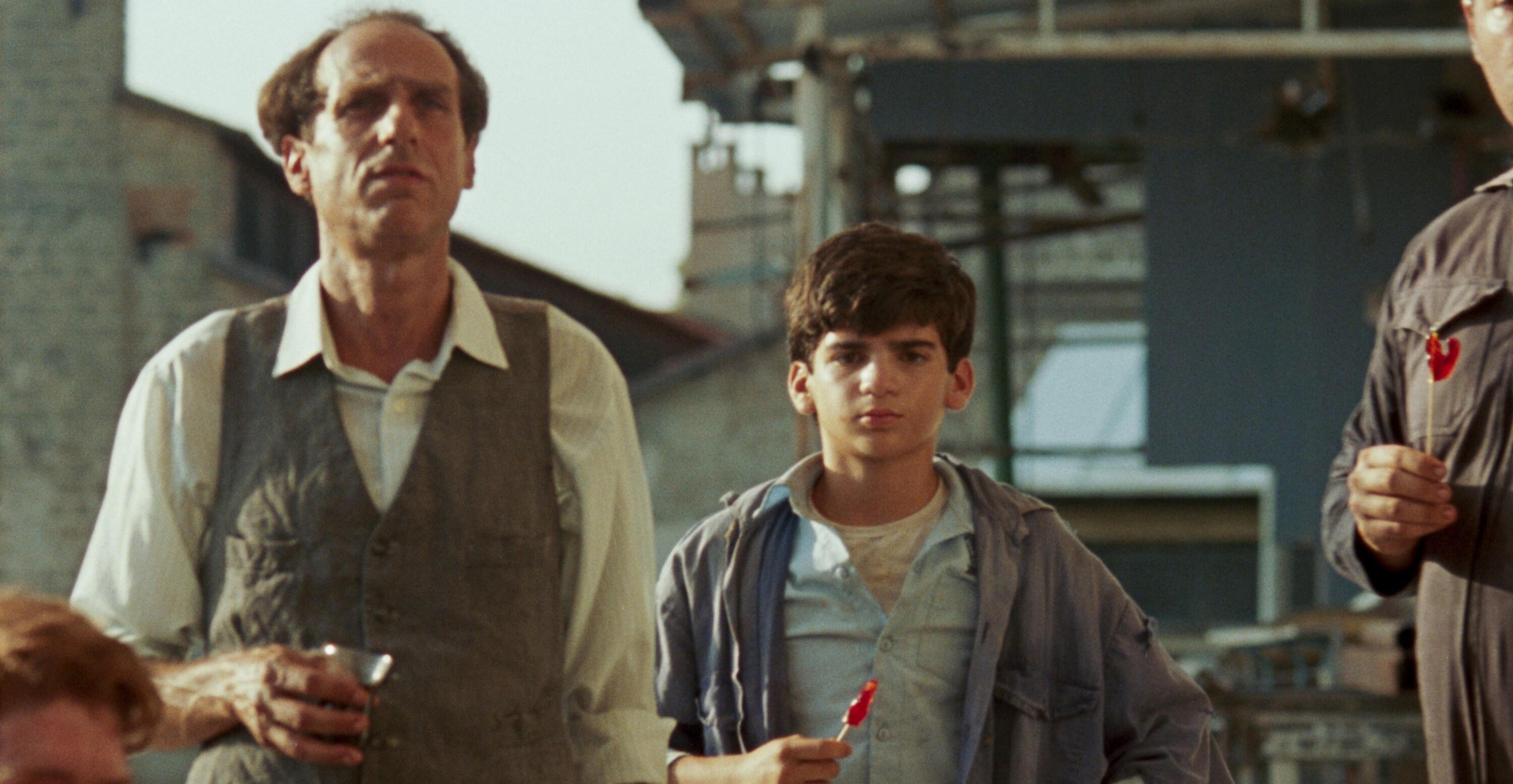 A young boy eating a lollipop staring forward while standing next to a balding man holding a glass in his hand in June Zero.