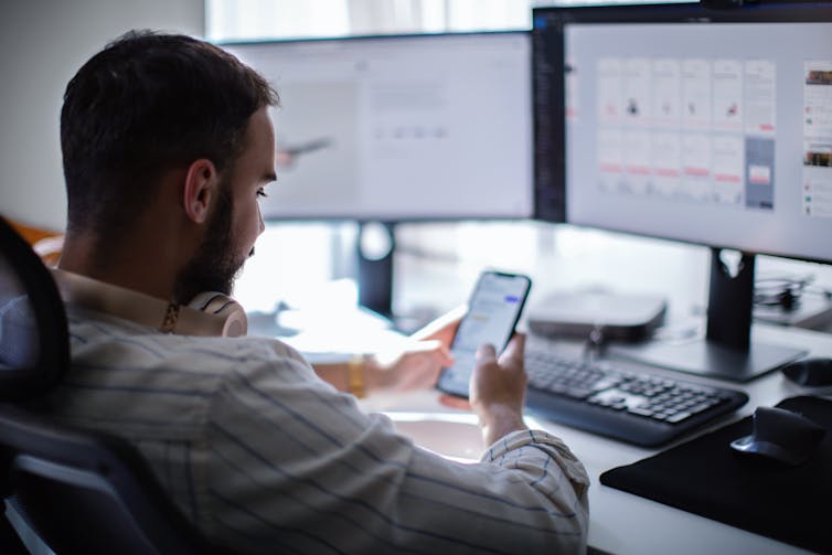 a man slouching in an office chair in front of a computer monitor looks at his phone