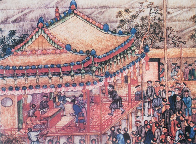 A painting showing people standing around a stage in a building which has tall pagodas.