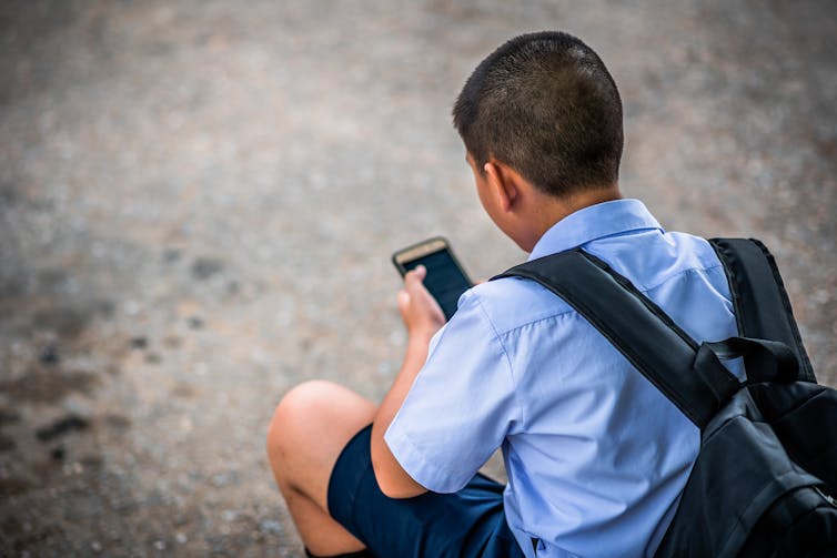 A teenage boy in school uniform and backpack looks at a phone.