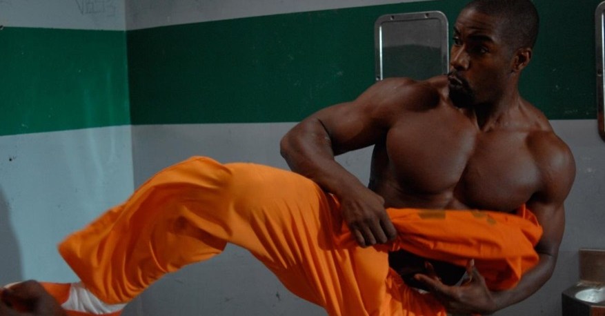 Michael Jai White, wearing prison orange, raises his leg to kick while also holding his shirt, ready to whip someone, in Blood and Bone