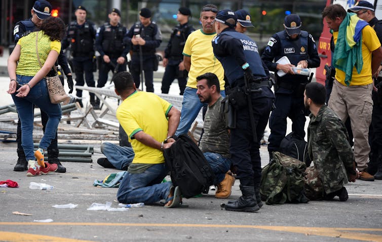 Men in restraints holding their arms behind their backs kneel on the floor with security guards around them.