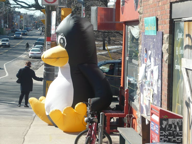 A street with a large inflatable penguin sitting down on the sidewalk.