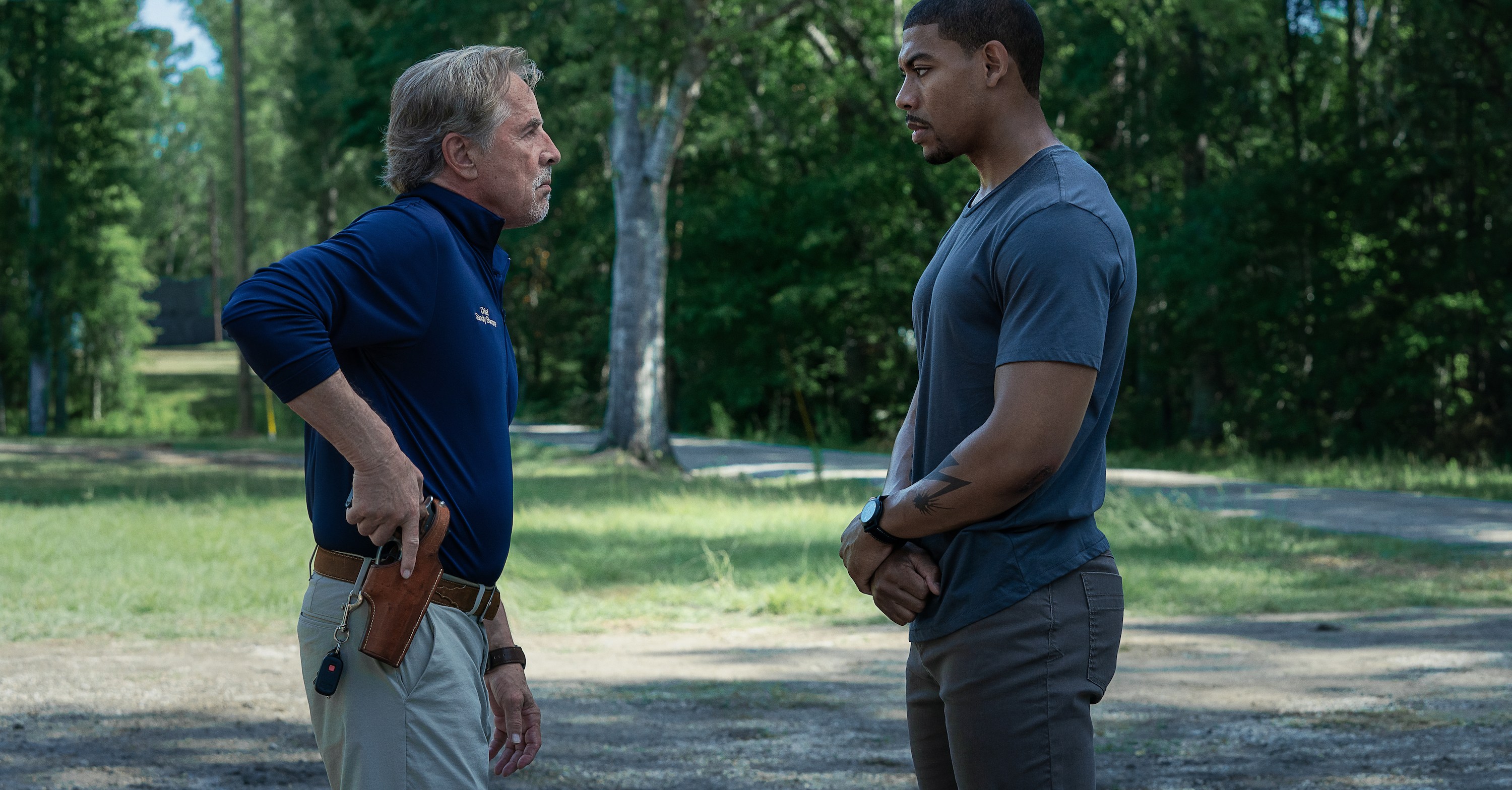 Police Chief Sandy Burnne (Don Johnson) puts a hand on his gun while standing outdoors, talking to visiting ex-Marine Terry Richmond (Aaron Pierre) in Jeremy Saulnier’s action movie Rebel Ridge 