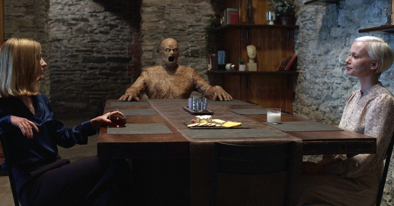 Two woman sitting across from one another at a table with a ghoulish wooden figure in Oddity.