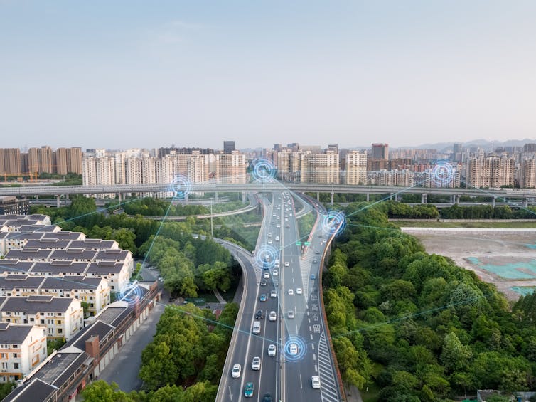 A cityscape with cars, some of which seem to be connected by thin blue lines, crossing a highway bridge over trees.